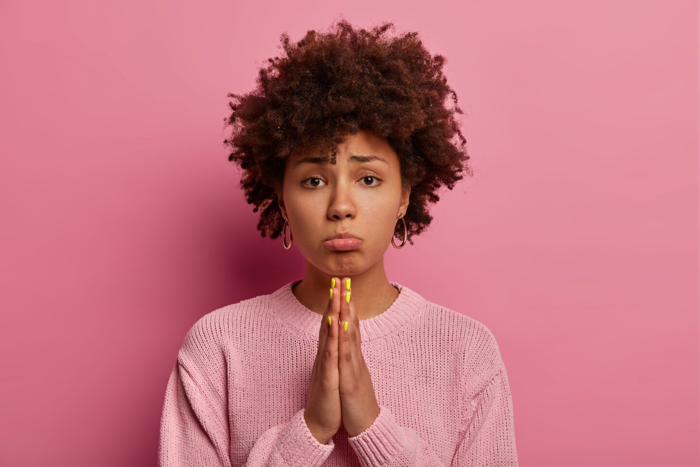 Hopeful sad uneasy curly woman begs for forgiveness, purses lips and keeps palms in praying gesture, asks mercy and says please, needs your help, wears casual jumper, poses over pink background