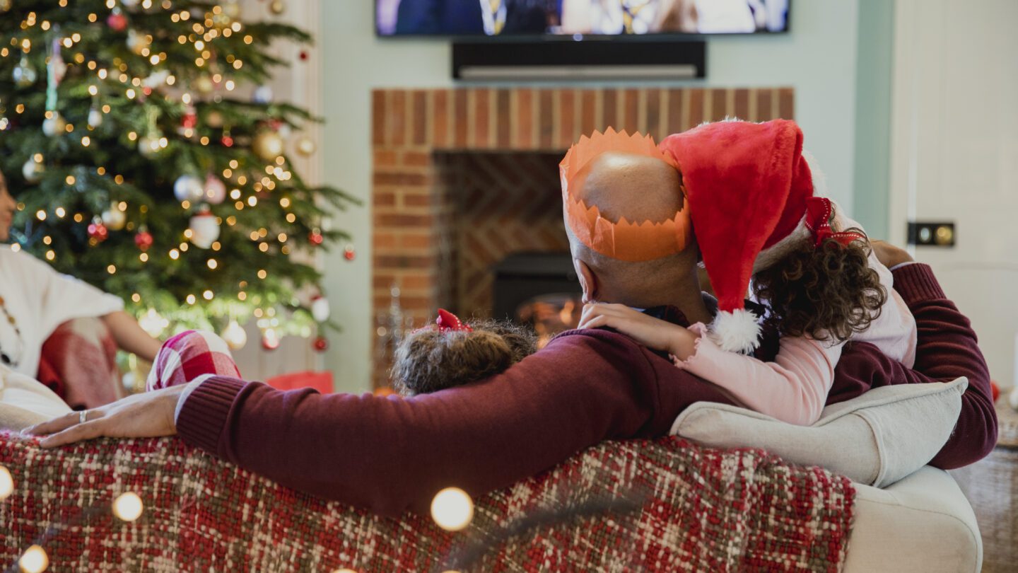 Father is curled up on the sofa ith his daughter at christmas time, watching television.