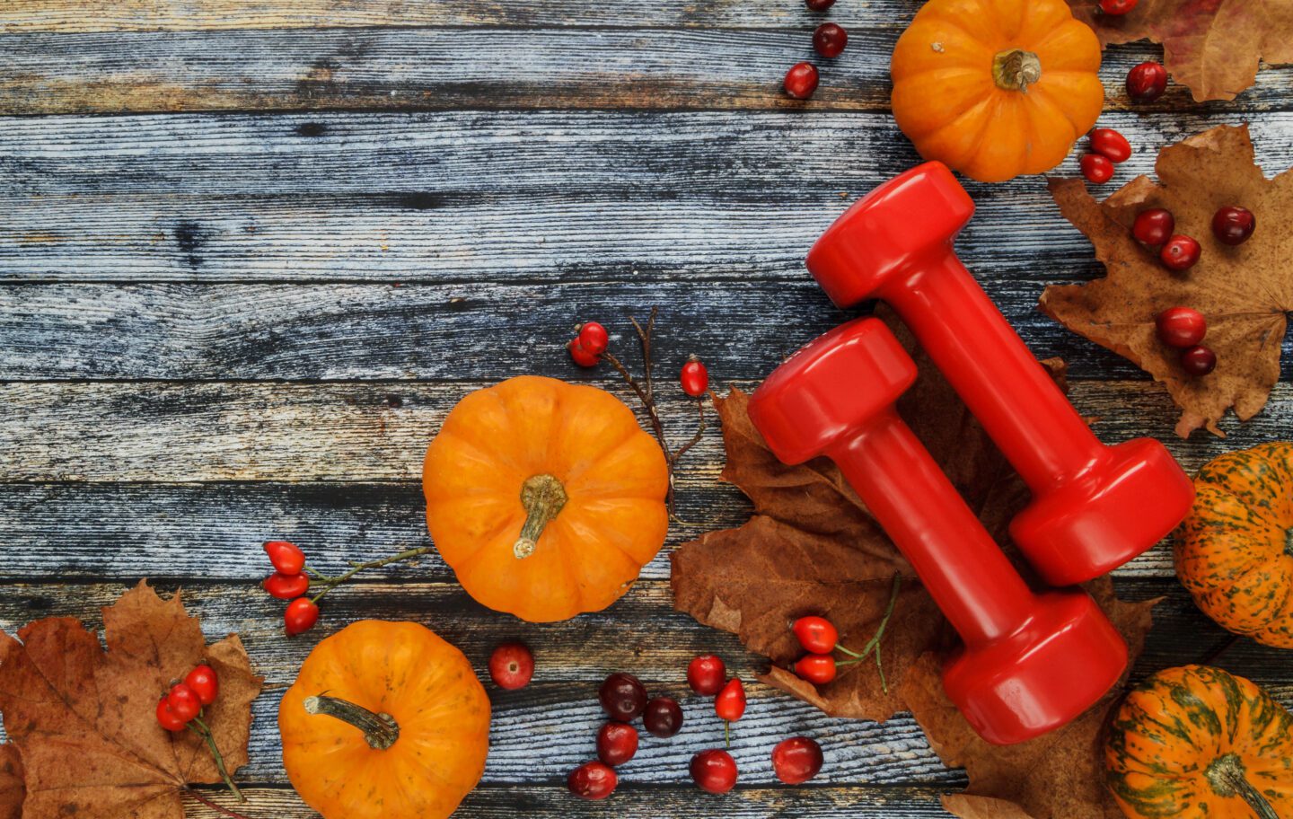 Two red dumbbells with pumpkins and autumn leaves. Healthy fitness lifestyle fall composition. Flat lay with copyspace on wooden background.