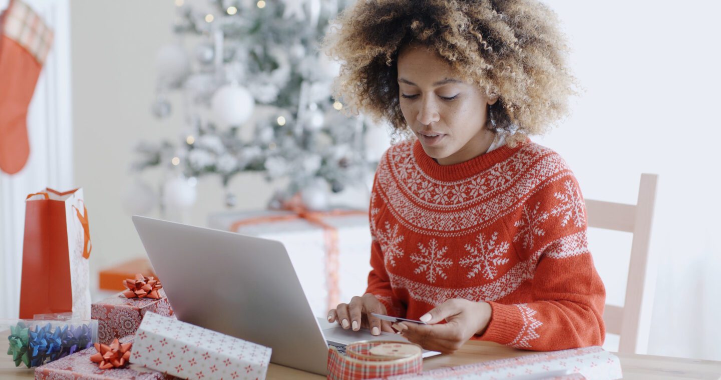 Young trendy African woman doing Xmas shopping online sitting in front of the decorated Christmas tree at home entering her card details on the computer